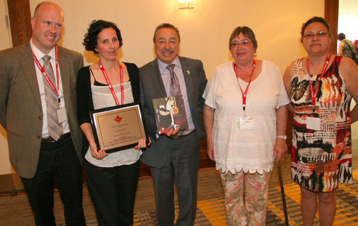Winner Julie Flett smilingly displays PMC Award Plaque, with Rob Smith, TNG to her left and Steve Beardy on her right joining in, along with two First Nation's Librarians.
